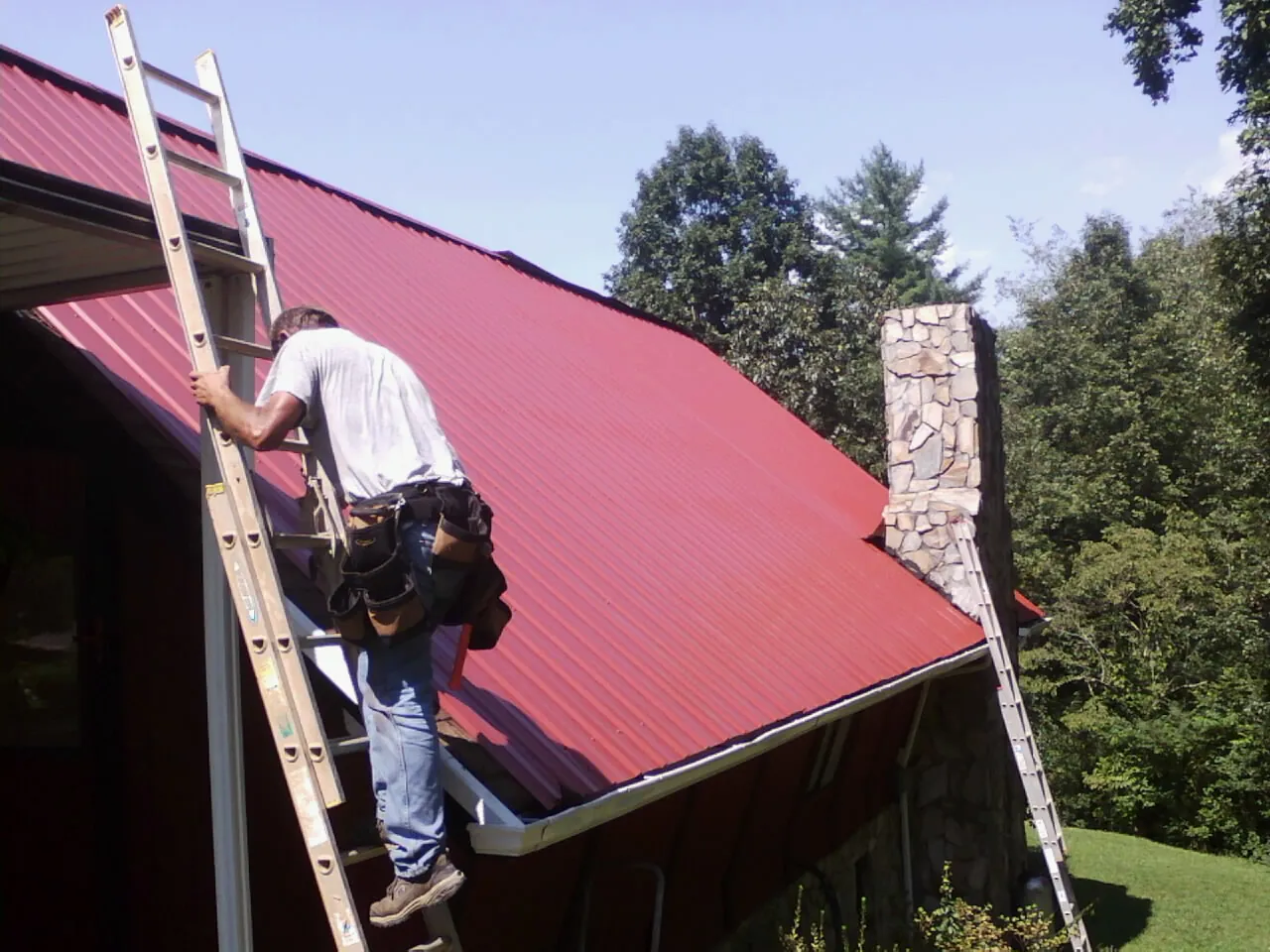 Roofer on a ladder beside a red metal roof and stone chimney — Soffit & Fascia Repair by Roofing Brothers in Moss Beach